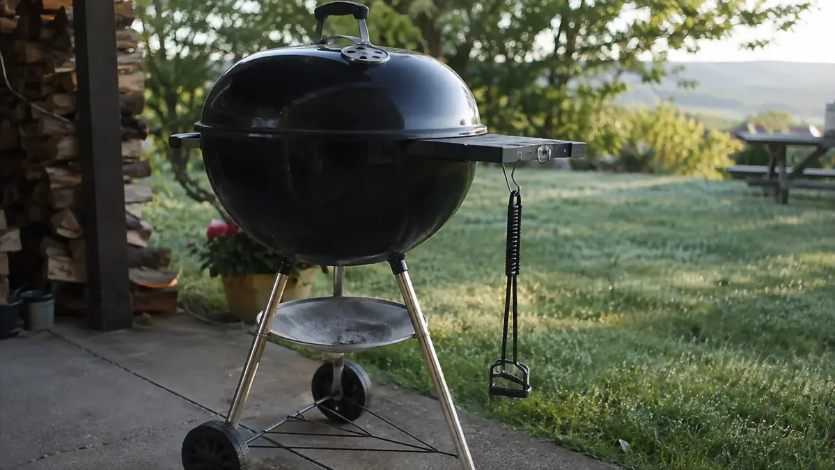 A cast iron grate lifter hanging on a hook beside a 22-inch charcoal kettle grill in a driveway, morning light