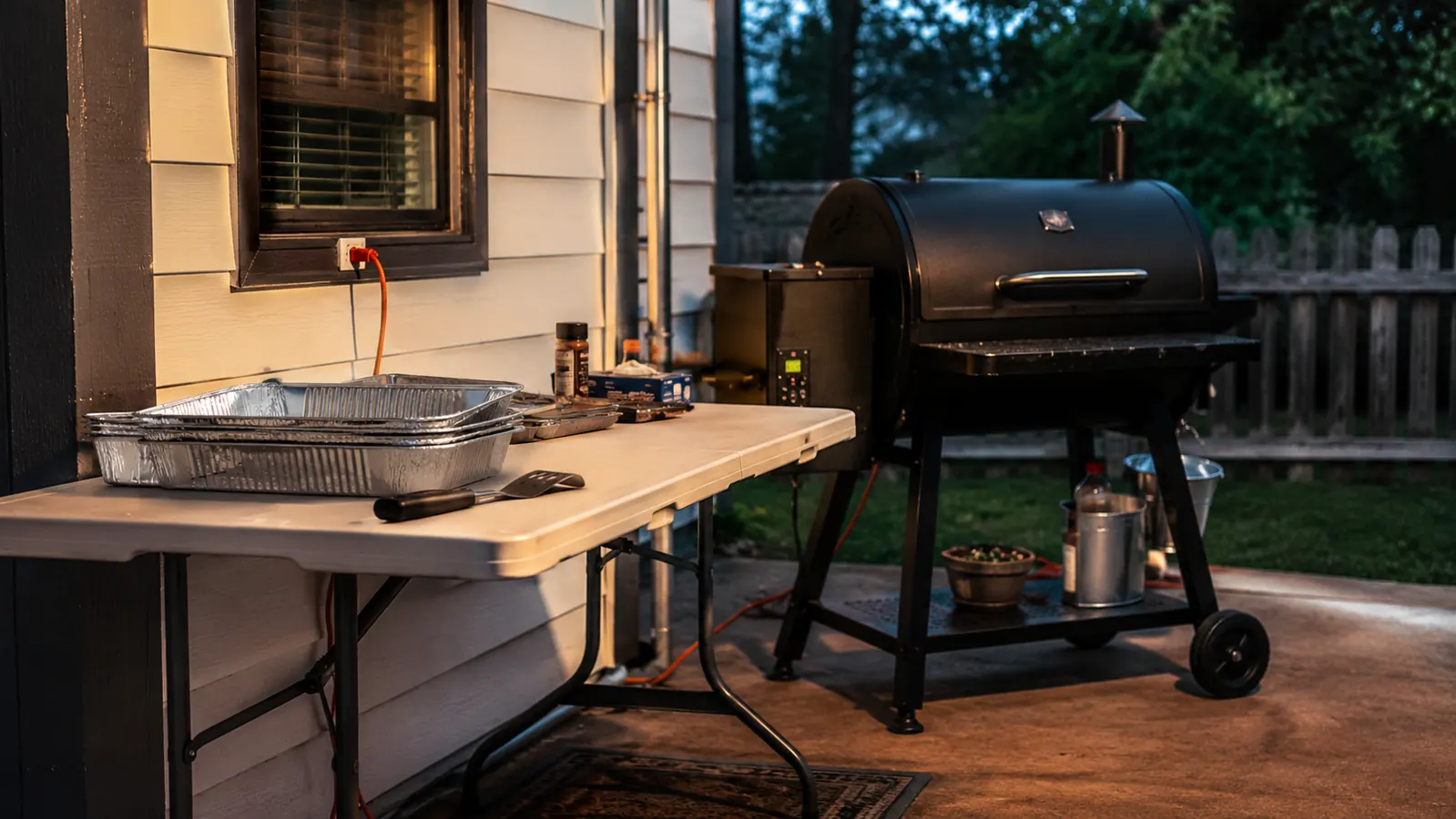 A folding table set up on a back patio next to a pellet grill near an electrical outlet