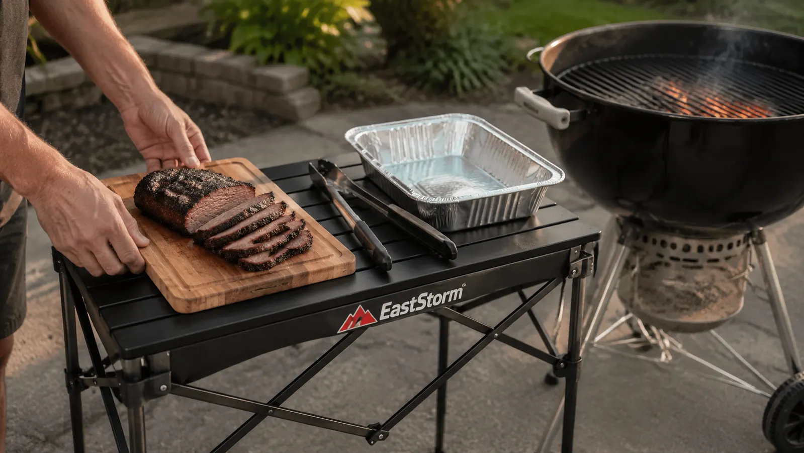 Hands setting a wooden cutting board with sliced meat onto a folding grill table beside a charcoal kettle grill
