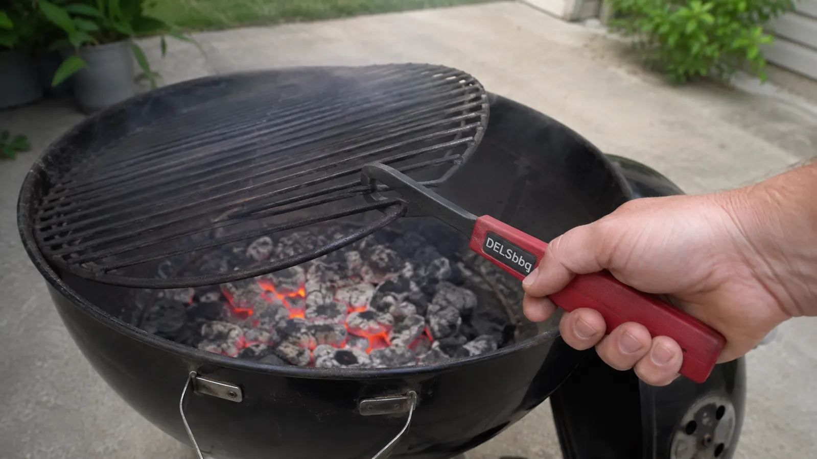 A hand using a cast iron grate lifter to hook and lift a hot cooking grate off a charcoal kettle grill