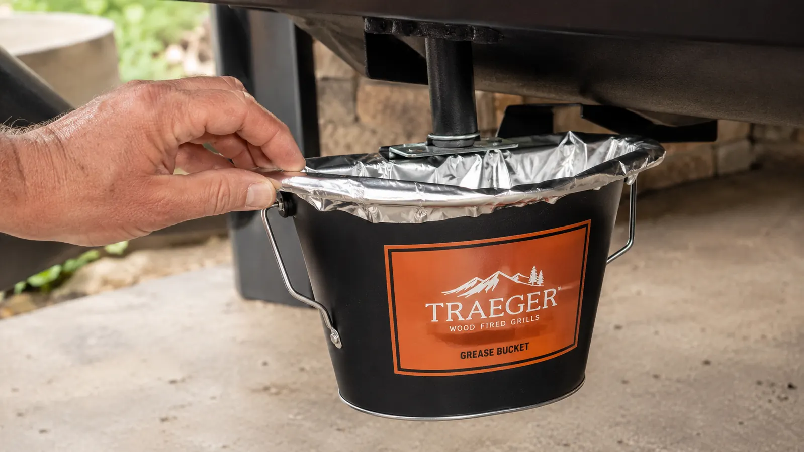 A hand folding a foil liner bag over the rim of a small metal bucket clipped to the side bracket of a pellet grill