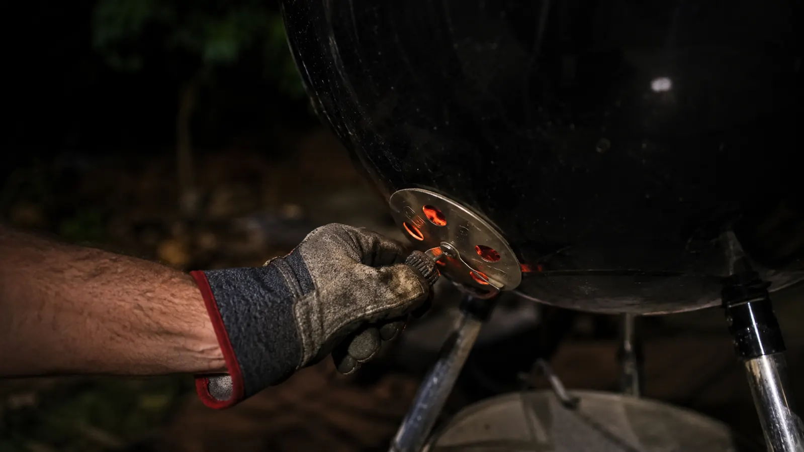 A gloved hand adjusting the bottom vent damper on a charcoal kettle grill by flashlight at night